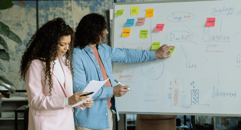 Equipe de duas pessoas em agência de marketing digital, apontando para um quadro branco com escrita e post itis de planejamento estratégico