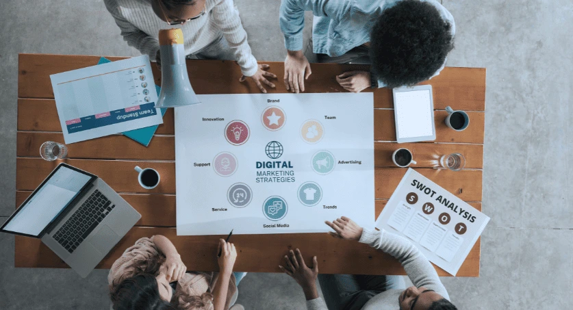 Equipe de uma agência de marketing digital sentadas em uma mesa de reunião com cartaz de planejamento estratégico ao centro, notbooks em volta e checklists de trabalho em papel