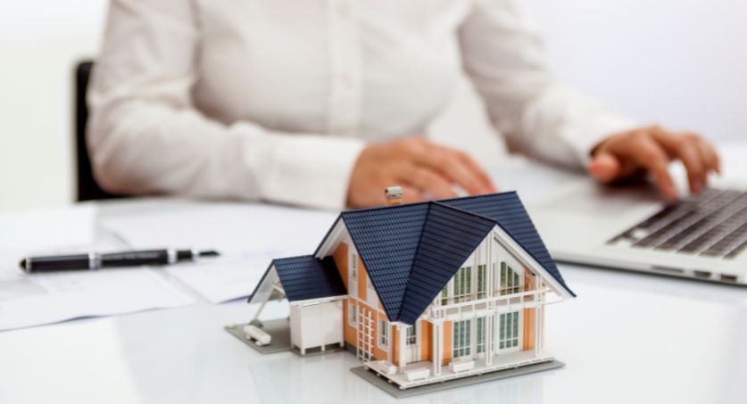 Homem no escritório de uma imobiliária com computador e miniaruta de casa de madeira na frente, analisando dados de investimento no marketing imobiliário