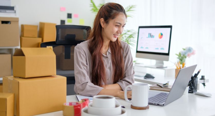 Mulher com caixas de papelão atrás e sentada em frente ao computador analisando gráficos para reposição de estoque sazonal