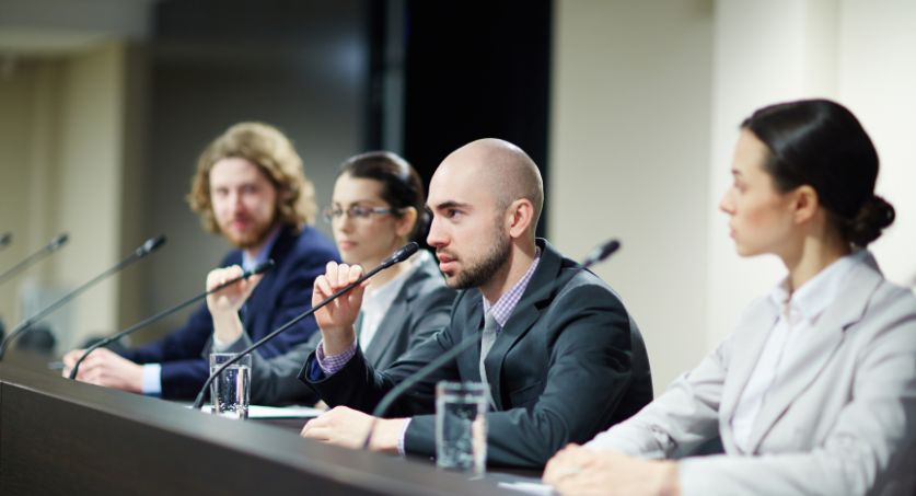 Reunião formal com profissionais sentados em bancada, utilizando microfones durante debate sobre marketing político e comunicação pública.