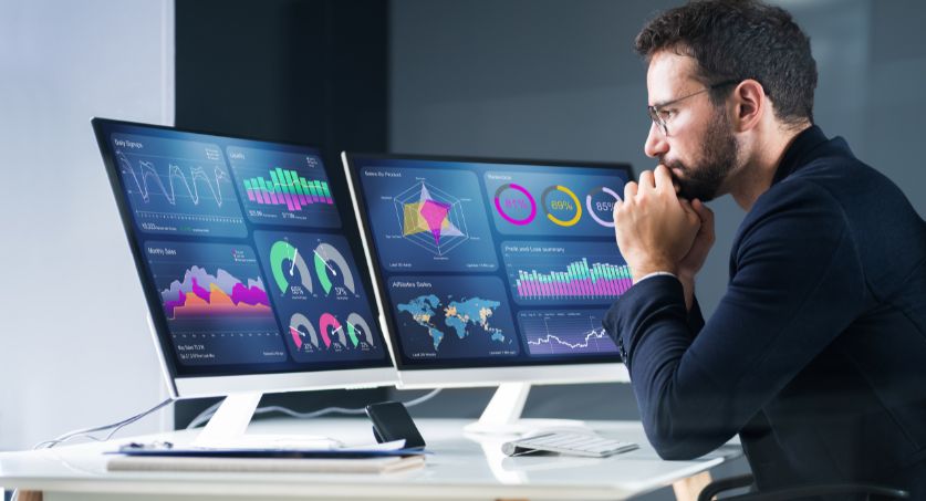 Profissional analisando dashboards de KPIs de marketing exibidos em dois monitores no escritório.