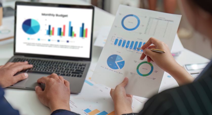 Equipe avaliando gráficos e relatórios de KPIs de marketing em notebook e documento impresso.